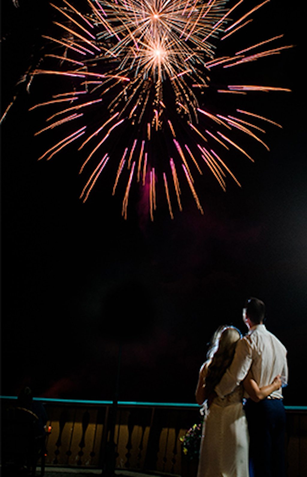 A newlywed couple embraces while they watch fireworks at their wedding.